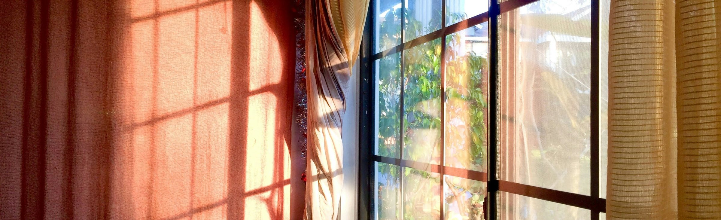 Warmes Licht fällt durch ein Fenster mit Vorhang, die Schatten der Fensterkreuze liegen auf rötlichem Stoff